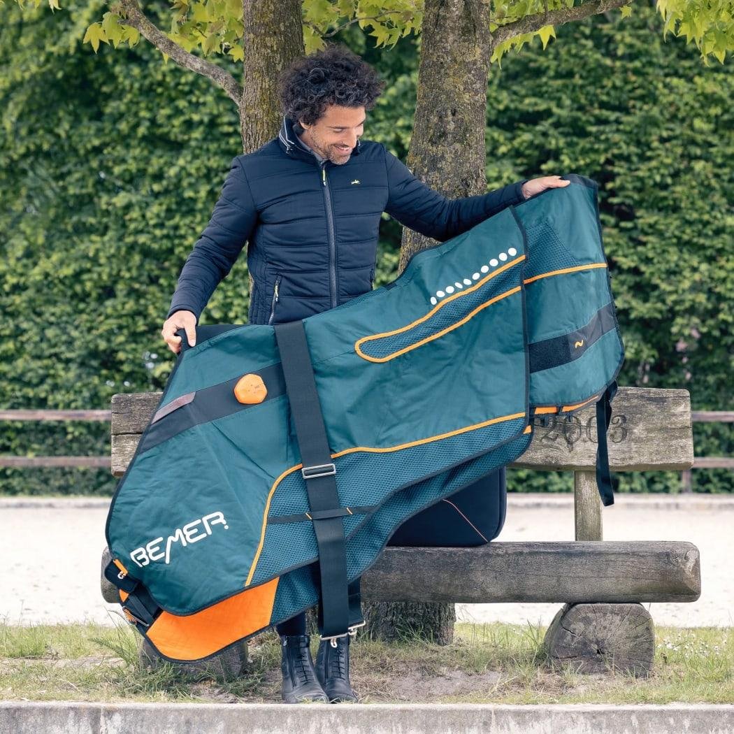A person holding up a teal BEMER horse blanket with orange trim beside a wooden bench outdoors